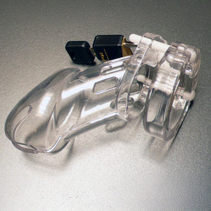 The assembled cage from the CB-6000 Package Male Chastity Kit is shown against a silver background. The cage is made of clear plastic with small cutouts and is shaped like a flaccid penis. It is attached to the base ring and locked with a small padlock.