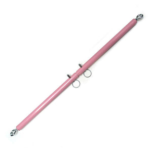 The Pink Adjustable Spreader Bar is displayed against a blank background. It is a long, metal, pink-colored bar with metal eyeholes at each end. It has two metal pegs in the middle that can be moved to adjust the length of the bar.