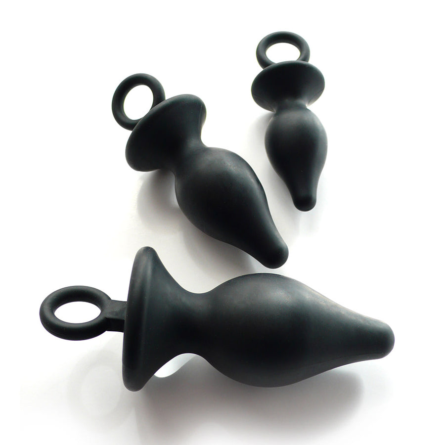 The Anal Trainer Kit is shown against a blank background. The kit has a small, medium, and large butt plug, all made of black rubber. The plugs are tapered with thin necks and wide bases at the bottom, and the bases have rings attached to them.
