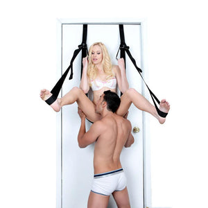 A blonde woman wearing a white bra sits against the door in the Door Jam Sex Sling, holding onto the handles at the top, with her feet in the stirrups. A dark-haired man is between her legs, licking the inside of her thigh.