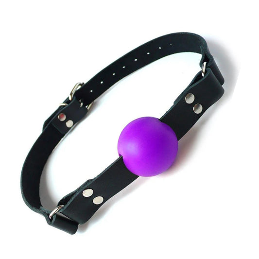 The purple rubber ball gag is displayed against a blank background. It is a matte purple rubber ball attached to a black leather strap with silver hardware, which buckles behind the head.