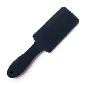 The Tantus Thwack Paddle is displayed against a blank background. The paddle is rectangular with a rounded handle and is made of black silicone.