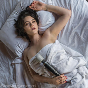 A nude woman with dark hair is shown wrapped in a white blanket laying on a bed with white sheets looking at the camera while holding a Doxy Vibrating Wand Massager in Black against her body.