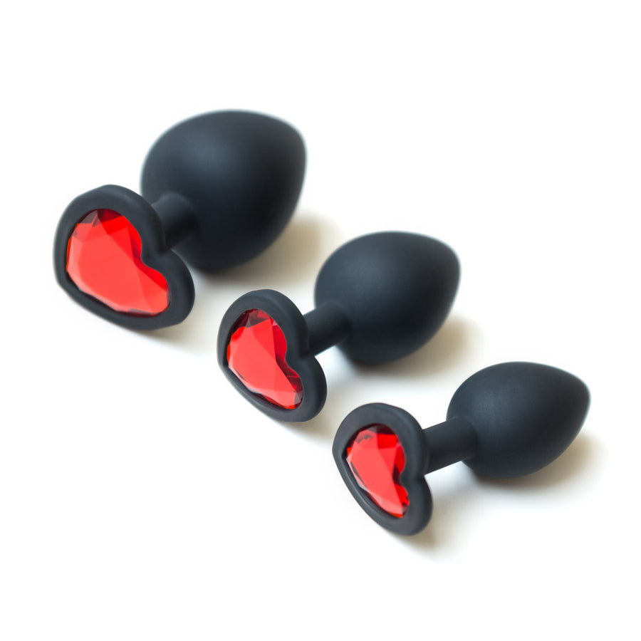 The 3 butt plugs from the Heart Anal Trainer Kit are arranged from smallest to largest against a blank background. They are made of black silicone and are tapered with thin necks and wide heart-shaped bases. They have a heart-shaped red gem on the bases.