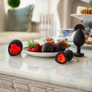 The black butt plugs with red gems from the Heart Anal Trainer Kit are arranged around a plate of chocolate-covered strawberries. They are on a white marble counter with various other plates of food in the background.