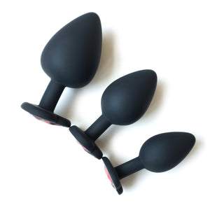 The three black butt plugs with red gems from the Heart Anal Trainer Kit are arranged from smallest to largest against a blank background.
