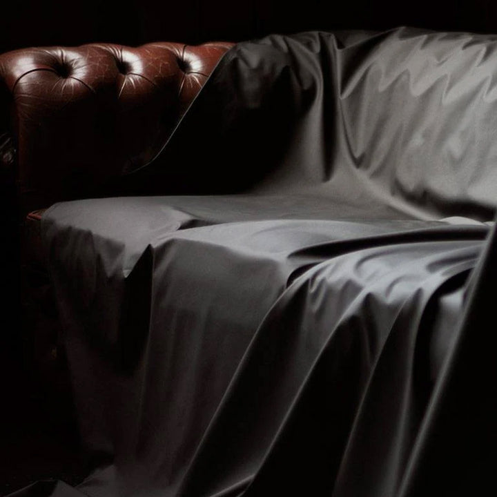 A brown leather couch is shown against a black background. All but the arm of the couch is covered by the Fluid Proof Protective Throw By Sheets Of San Francisco, a black sheet that drapes onto the floor. On the floor is a knocked-over glass on the sheet.