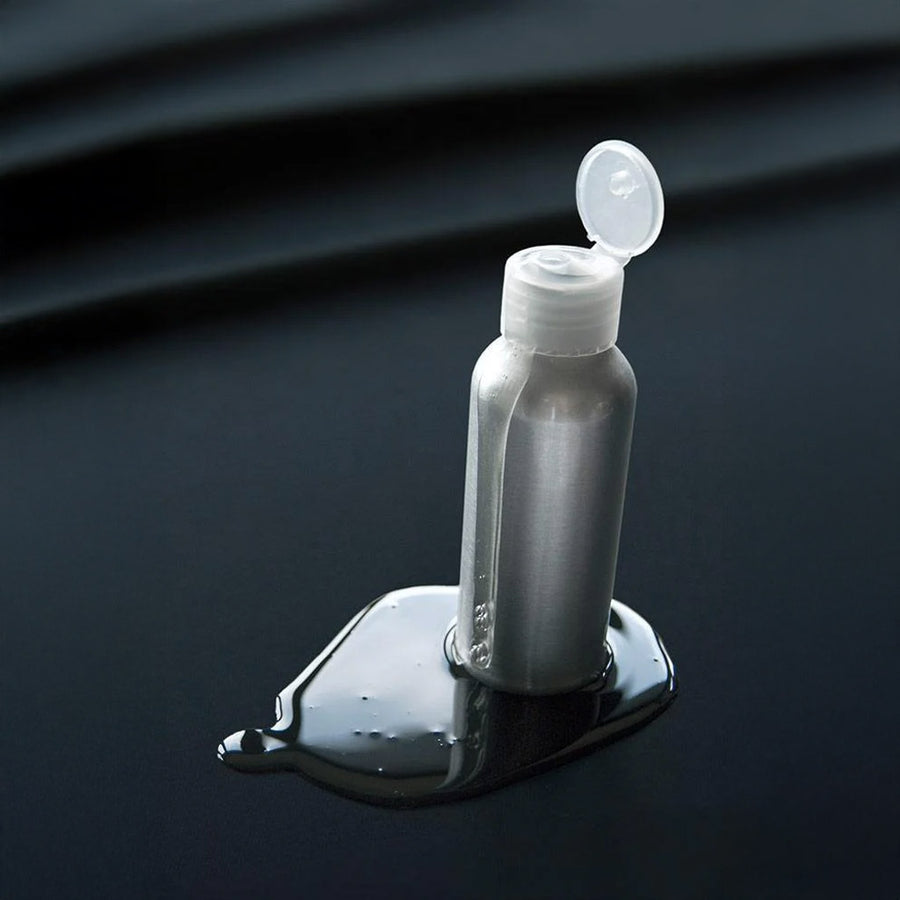 A silver bottle of lube with a flip-cap is shown standing on the Fluid Proof Protective Throw By Sheets Of San Francisco. There is a pool of lube under the bottle, but the fluid rests on top of the sheet instead of soaking into it.