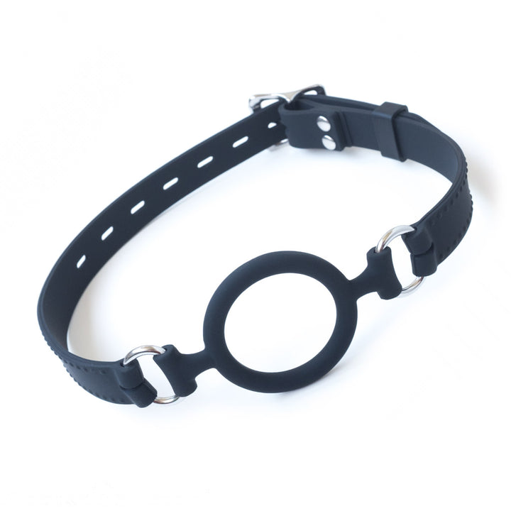 The black Silicone O-Ring Bondage Mouth Gag is shown against a blank background. It has a silicone O-ring in the center connected to metal O-rings, which attach the gag to its strap. The strap is made of black silicone with a faux-stitched border.