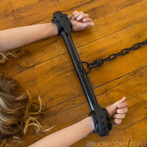 A close-up of a woman’s wrists is shown against a wooden floor. Her wrists are locked in the black Big Barrel 12 Inch Wrist Spreader Bar With black buckling Cuffs. A heavy black chain is connected to a D-ring in the middle of the spreader bar.