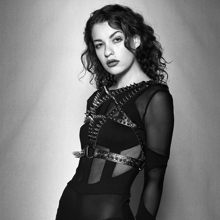 A black and white photo of a brunette, curly-haired woman posing in front of a backdrop. She wears a mesh shirt with the black leather Spiked Pentagram Bust Harness on top. The harness has straps arranged in the shape of a pentagram over her chest.