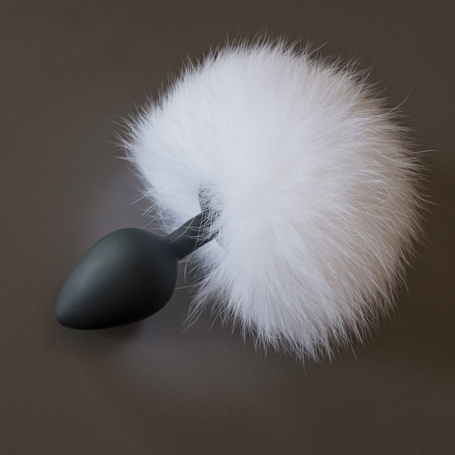 The White Rabbit Fur Bunny Tail Silicone Butt Plug is displayed against a gray background. The plug is made of matte black silicone and is small and tapered with a thin neck and wide base. Attached to the base is a large puff of white fur.