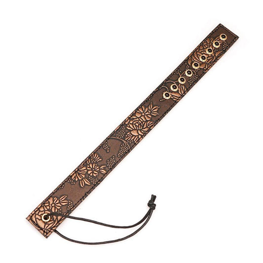 The Vegan Floral Ruler With Gems is shown against a blank background. It is a piece of brown faux leather shaped like a ruler with a floral design in light brown. There is a row of black gems at the top of the ruler and a wrist loop at the bottom.