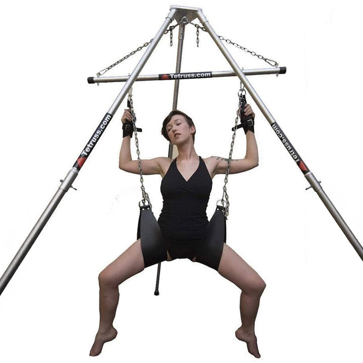 The Tetruss Portable Dungeon Deluxe Bundle is shown in use against a blank background. The spreader bar is connected to the frame, supporting a woman sitting in the leather slings. Her suspension wrist cuffs are attached to the spreader bar.