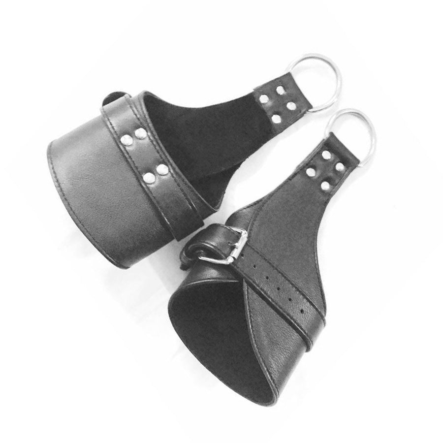 The Tetruss Wrist Or Ankle Suspension Cuffs are shown against a blank background. They are shaped like typical cuffs, but one side has an extended piece of leather with an O-ring attached to it. They are made of black leather with silver hardware.