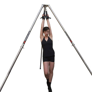 A woman wearing black shorts and a tank top is shown standing underneath the Tetruss Suspension Bondage Frame. She is wearing the Tetruss Wrist Or Ankle Suspension Cuffs on her wrists, which are attached to the top of the frame.