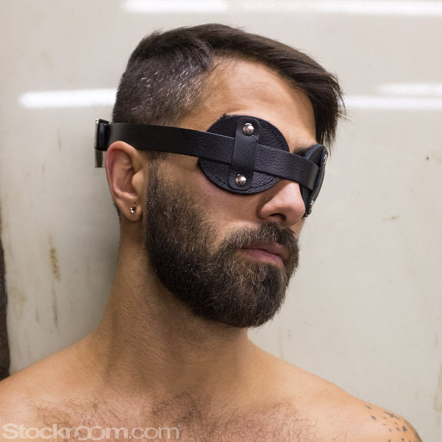 A closeup of a dark-haired man’s face with facial hair is shown. He is standing against a blank wall and wearing the Adjustable Aviator Blindfold, which is black leather and resembles aviator goggles.