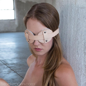 A blonde woman is shown from the shoulders upwards, sitting against a concrete wall. She wears the Institutional Fleece Lined Blindfold, which is made of tan leather and shaped like aviator goggles.