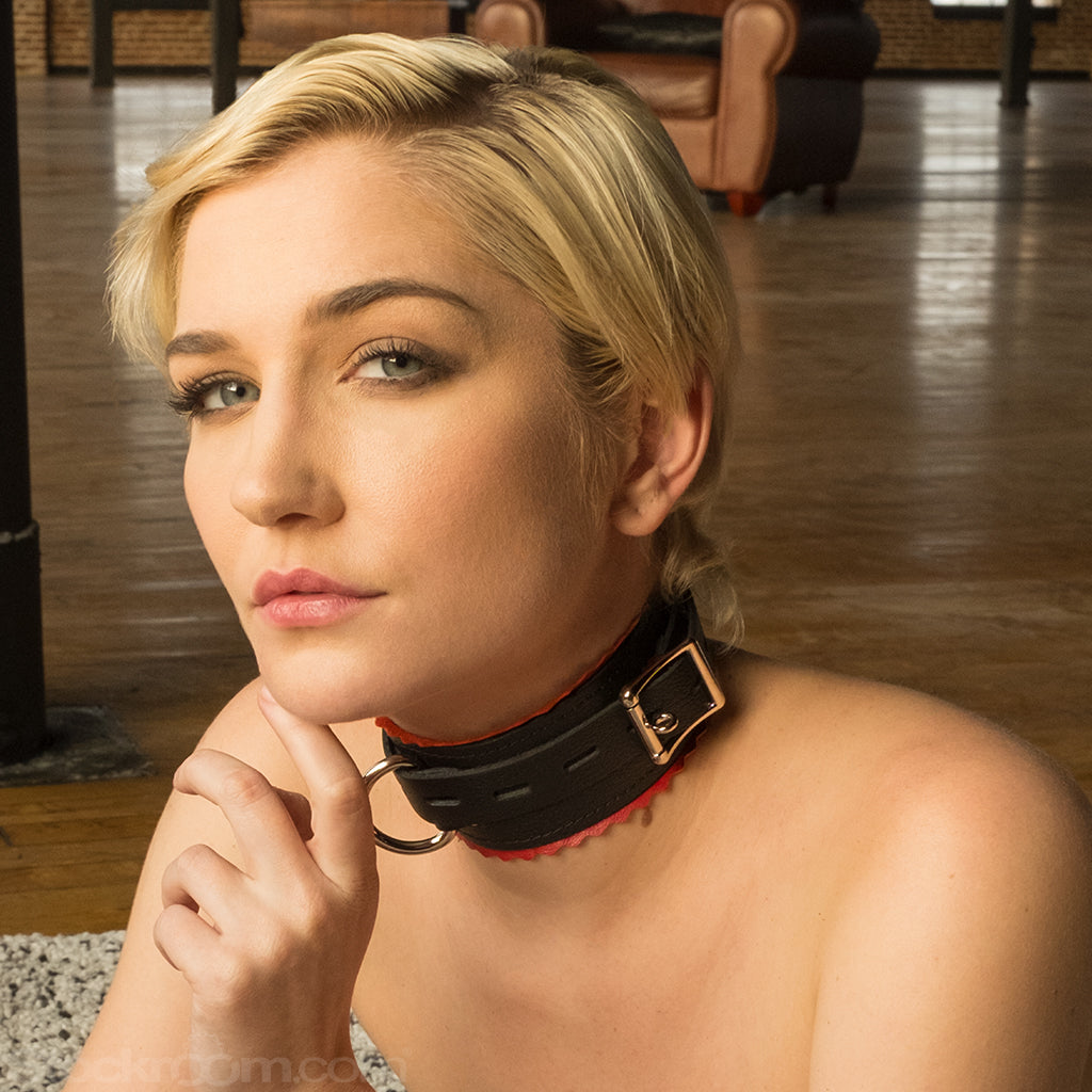 A close-up of a blonde woman wearing the Leather Buckling Collar with Scalloped Edges is shown. The collar is made of a wide piece of black leather with a smaller notched strip wrapped around it. It has red scalloped trim and metal hardware.