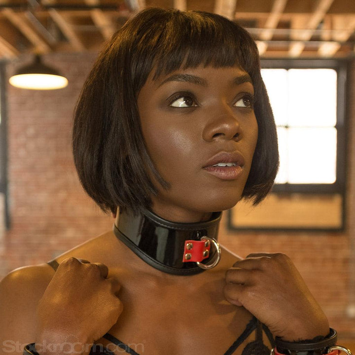A close-up shows a woman with a black bob from the shoulders upwards. She wears the Firecracker Patent Leather Posture Collar, which is a small black patent leather posture collar with red accents.