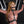 Load image into Gallery viewer, A woman with red hair wearing a mauve latex catsuit stands against a black St. Andrew’s Cross in front of a black wall. Her wrists are cuffed above her, and she has a black bit gag in her mouth. Over her catsuit, she wears the black PVC Bust Harness.
