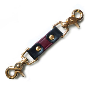 The JT Signature Collection Restraint Clip is shown against a blank background. The clip is a short piece of Bordeaux leather with black accents. Each end has a gold claw-style clip, and the hardware securing the clips is gold as well.