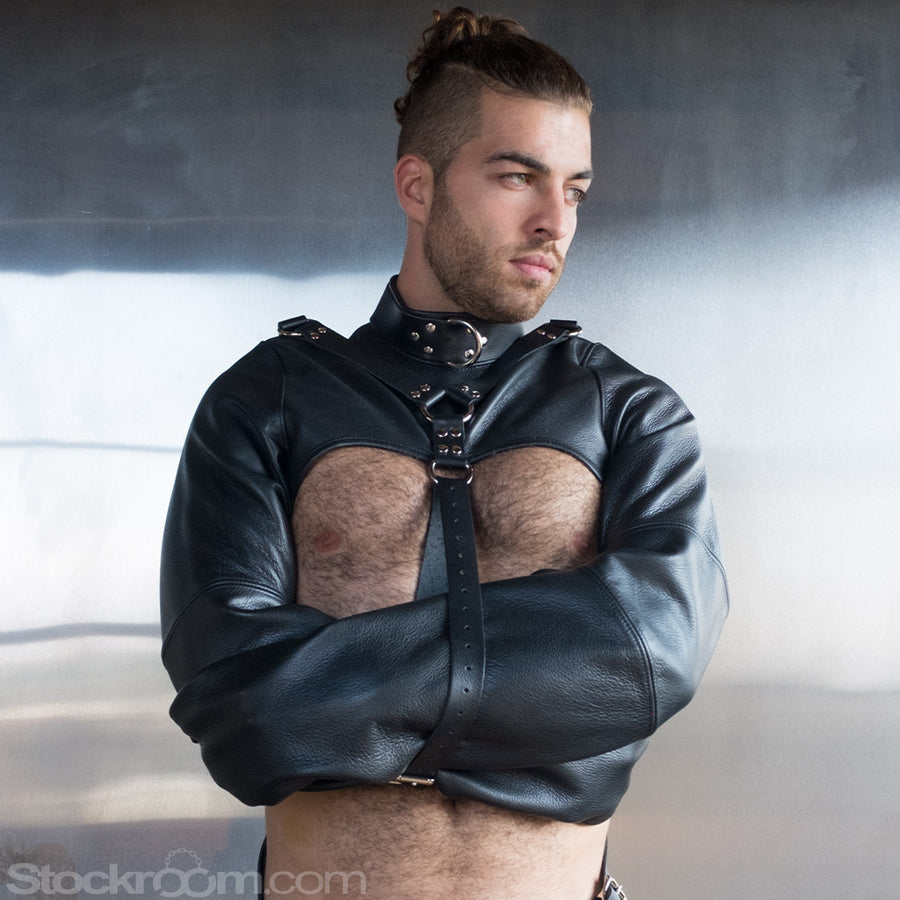 A man with dark, pulled-back hair and chest hair stands in front of a metal wall, wearing the Men's Bolero Straitjacket. The jacket is black leather and cuts off above his pecs. His arms are crossed and encased in the sleeves.