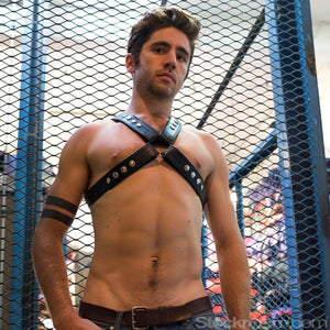 A shirtless brunette man in jeans stands in front of a mesh chain fence. He wears the Garment Leather Chest Harness, made of black leather with silver hardware. The harness is shaped like an X, with straps going over his shoulders and under his arms.