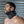 Load image into Gallery viewer, A brunette man with short facial hair is shown from the shoulders up, standing in front of a metal wall. He wears the black Deluxe Padded Leather Posture Collar, which covers his entire neck and has a projection that supports his chin.
