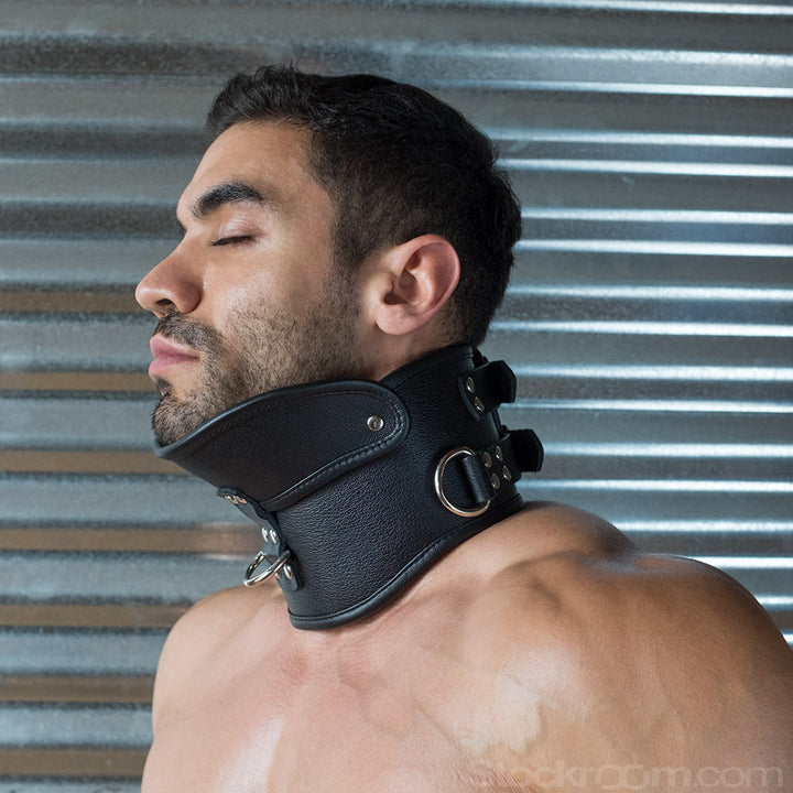 A brunette man with short facial hair is shown from the shoulders up, standing in front of a metal wall. He wears the black Deluxe Padded Leather Posture Collar, which covers his entire neck and has a projection that supports his chin.