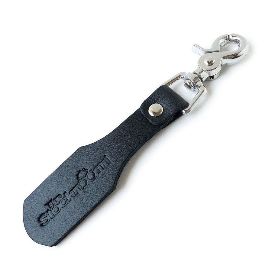 The Stockroom Branded Leather Keychain Paddle is shown against a blank background. They keychain is a small piece of black leather shaped like a paddle, attached to a metal snap hook, with The Stockroom logo branded onto it. 