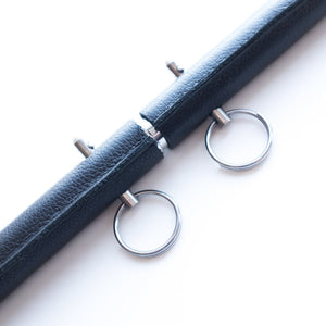 A close-up of the pins in the black Leather Wrap General Purpose Spreader Bar is shown against a blank background. The pins are small metal rods with rings on top. There is a small separation in the leather in the middle of the bar where it expands.