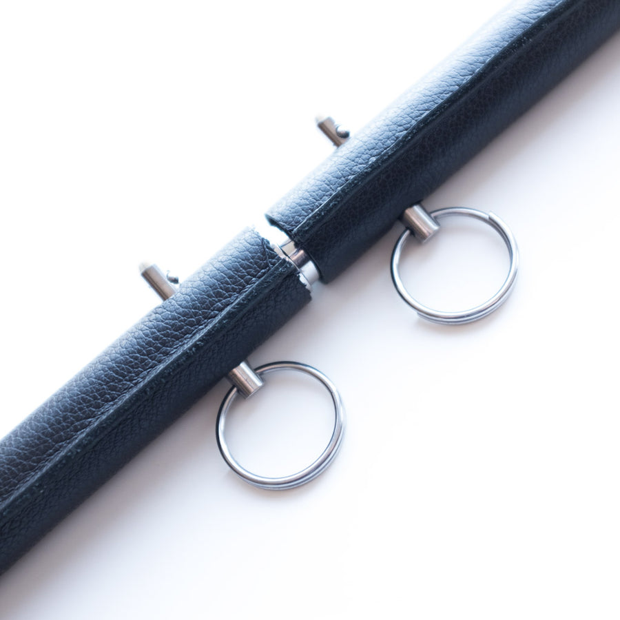 A close-up of the pins in the black Leather Wrap General Purpose Spreader Bar is shown against a blank background. The pins are small metal rods with rings on top. There is a small separation in the leather in the middle of the bar where it expands.