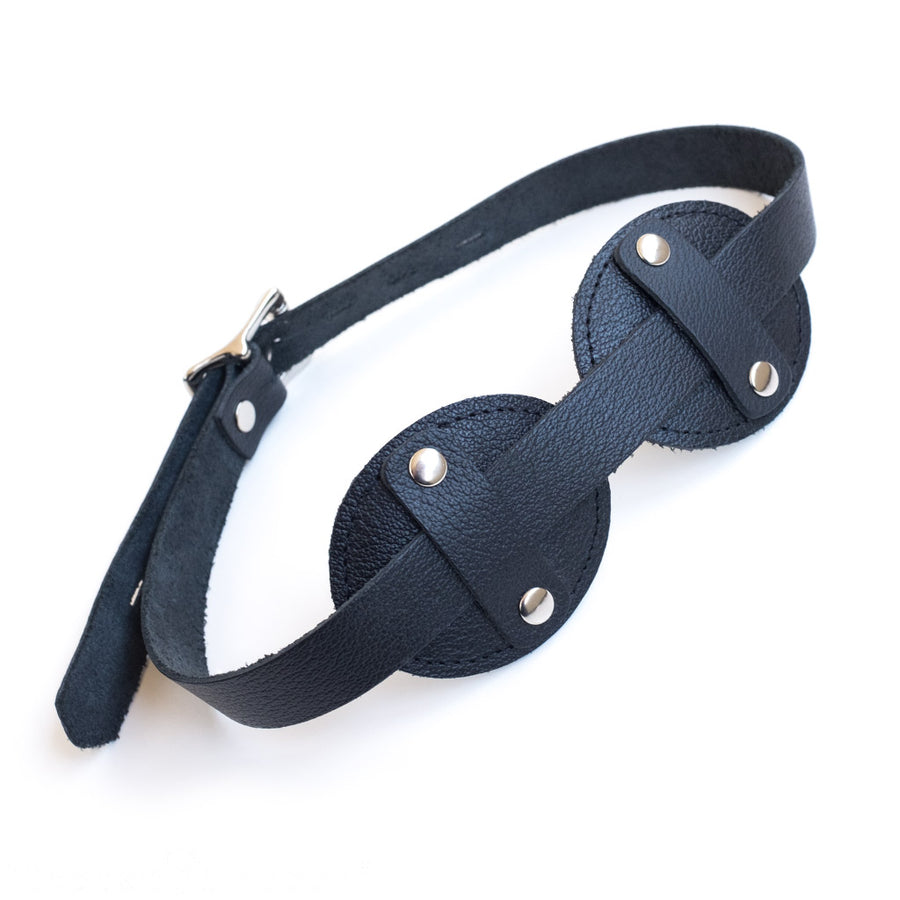 The Foam Padded Aviator Blindfold is shown against a black background. It is shaped like aviator goggles. The strap is a strip of leather that runs across the goggles, held in place by a small vertical piece of leather on each eye, secured by rivets.