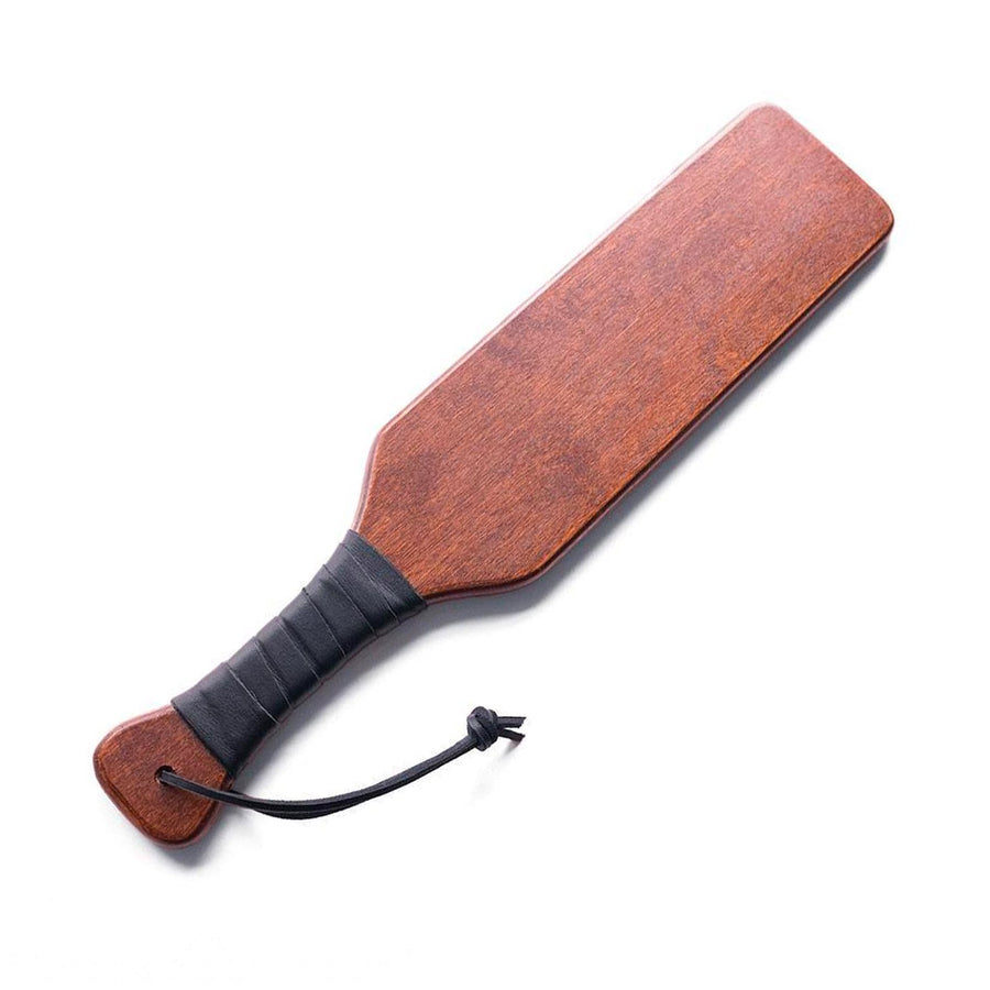 The Leather Wrapped Wood Spanking Paddle is displayed against a black background. The paddle is rectangular and made of dark, smooth wood. The handle of the paddle is wrapped in a black leather strip. A wrist strap is attached to the bottom of the paddle.