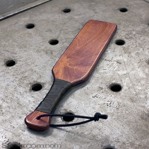 The Leather Wrapped Wood Spanking Paddle is laid out on a metal surface.