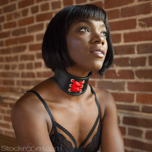 A woman with a black bob wearing black lingerie kneels in front of a brick wall. She wears the Red Laced BDSM Posture Collar, a small posture collar made of black leather with decorative red corset lacing on the front.