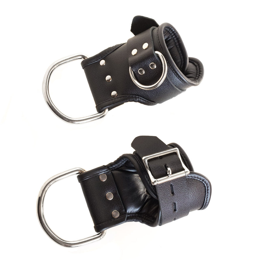 The Padded Suspension Cuffs, made of black leather, are displayed against a blank background. The top of the cuffs extends lower on one side. This side has a large silver D-ring attached to it. The cuffs also have a D-ring around the wrists.