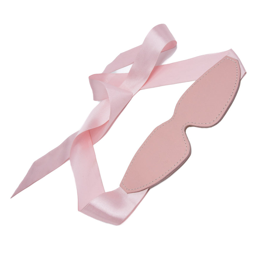 The Stupid Cute Blindfold is displayed against a blank background. It is made of light pink leather with white stitching along the border. It has two pink ribbons that can be tied in the back to secure it.