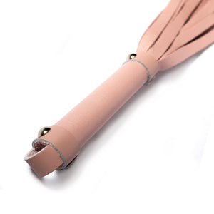 A close-up of the Stupid Cute Baby Pink Leather Flogger handle is shown against a blank background, displaying the small leather loop at the base of the handle and the metal hardware.