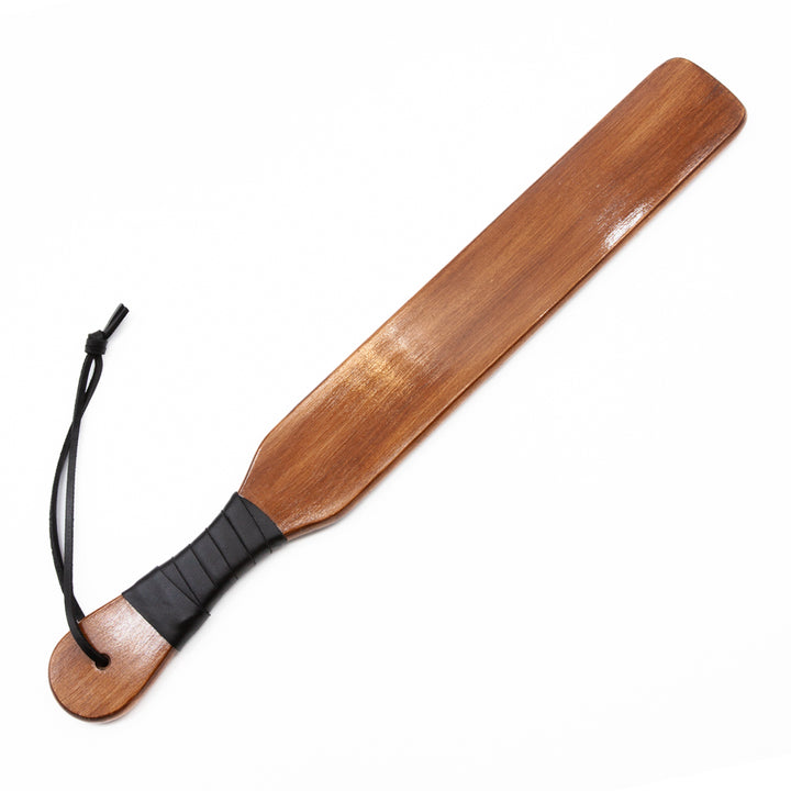 The Leather Wrapped Narrow Wood Spanking Paddle is shown against a blank background. The paddle is made of light-colored wood in the shape of a rectangle with a thinner handle. The handle is wrapped in black leather and has a wrist loop.