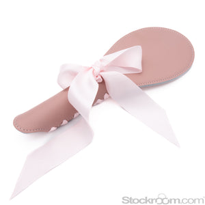 The Stupid Cute Pink Leather BDSM Spanking Paddle is shown against a blank background. It is shaped like a ping-pong paddle and has accent stitching around the border. It has a light pink ribbon laced up the back of the handle and tied in a bow.