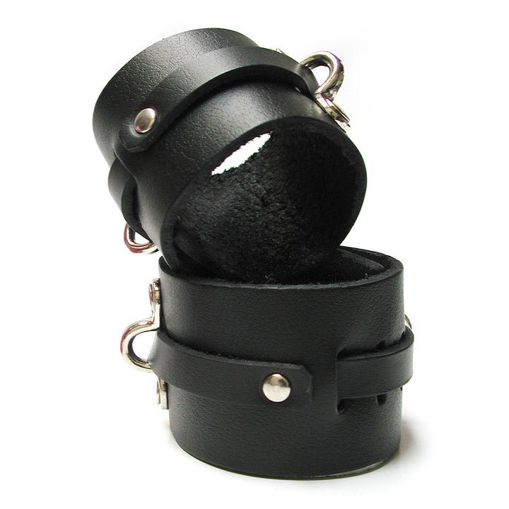 The KinkLab Bondage Basics Leather Wrist Cuffs are shown against a blank background. They are made of a wide piece of black leather with a thin piece of leather that wraps around them, through notches and under metal D-rings.