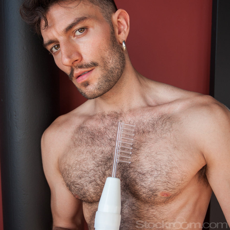 A shirtless man with dark hair, facial hair, and chest hair stands against a red and black background. He holds the wand with the electrode comb from the Kinklab Neon Wand® Electrosex Kit.