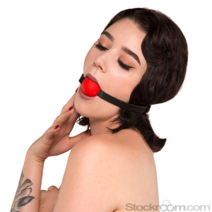 A woman with shoulder-length curly dark hair is shown against a blank background, wearing the Kinklab Red Silicone Ball Gag With A Leather Strap, which is wrapped around her head over her hair.