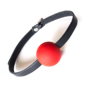 The Kinklab Red Silicone Ball Gag With A Leather Strap is displayed against a blank background. The gag is a ball made of matte red silicone, which has a thin leather strip on each side that buckle in the back.