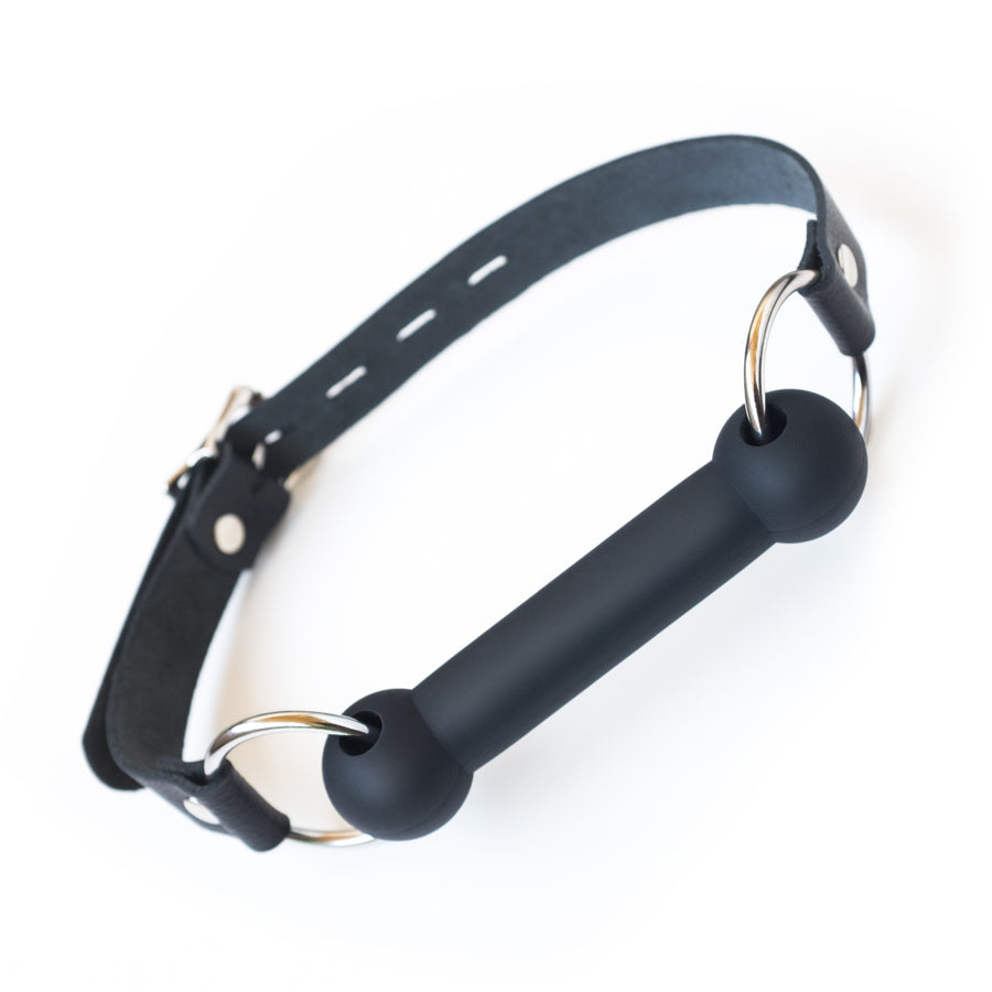 The Kinklab Silicone Bit Gag With A Leather Strap is shown against a blank background. The gag bit is made of matte black silicone and attached to the leather head strap with metal O-rings. 