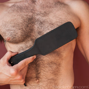 A shirtless man with brown chest hair is shown standing against a red background. He is holding the Kinklab Thunderclap Electro Conductive Paddle Neon Wand® Attachment in front of his chest.