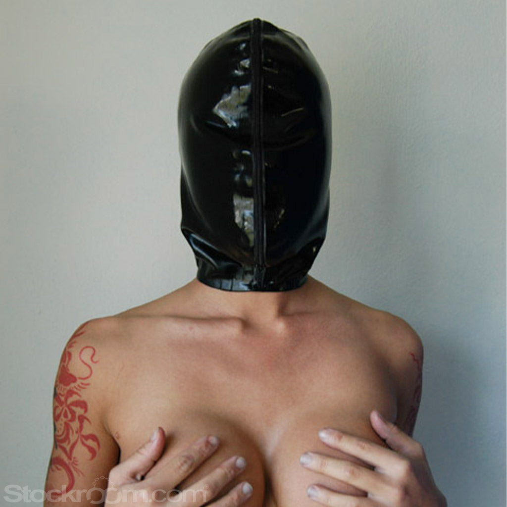 A topless woman is shown from the waist up, standing against a wall and covering her breasts with her hands. She wears the Extreme Rubber Hood zipped up, completely covering her face.