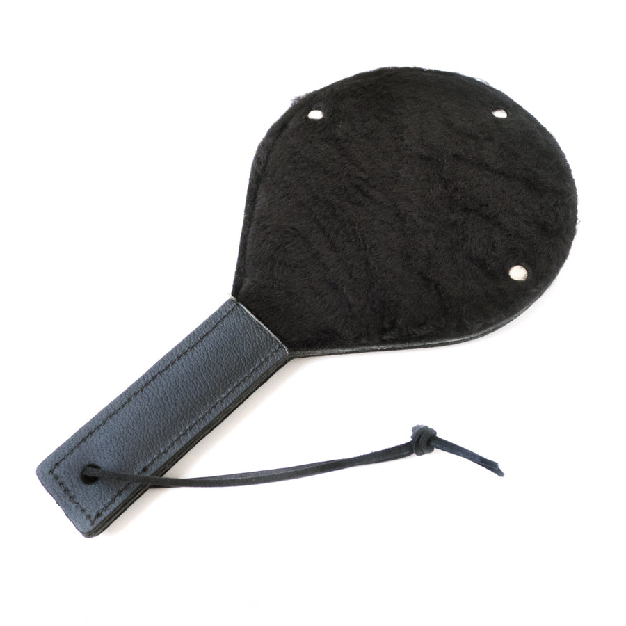 The Black Leather Gentle Persuasions Paddle is shown against a blank background with the fuzzy side facing up. The paddle is rounded with a straight leather handle with a wrist loop at the base. The paddle is covered in black faux fur.